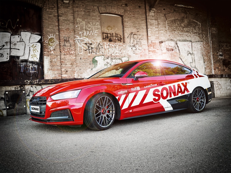 Blank och polerad Audi med SONAX-logga på sidan står uppställd framför industribyggnad